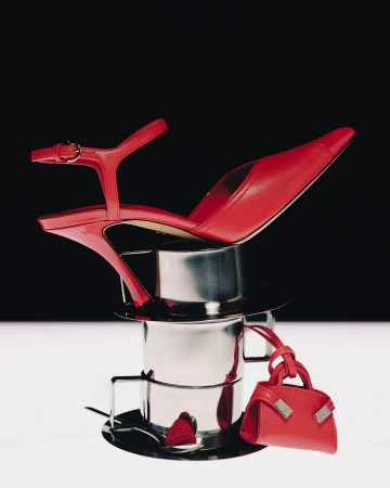 Red high heel shoe and miniature red handbag balanced on a stack of metal cups against a two-tone black and white background.