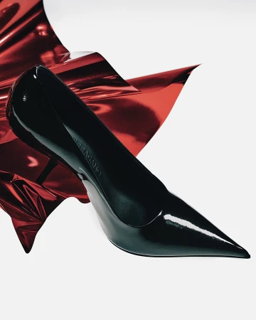 Black patent leather stiletto pump resting against a crumpled metallic red sheet on a white background