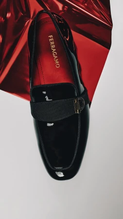Top-down view of a black patent leather loafer with a gold buckle resting on a red metallic foil and white background.