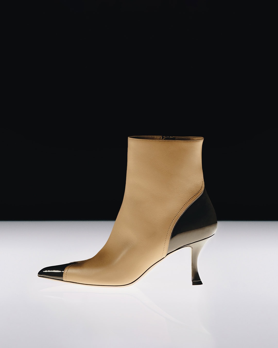 Tan leather ankle boot with black patent toe and heel cap on a reflective white surface against a dark background