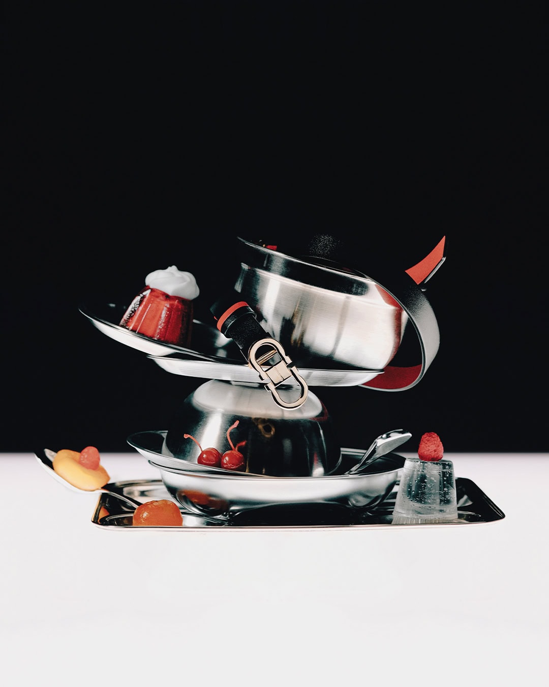 Stacked metallic cookware and trays garnished with desserts and cherries against a dark and light background