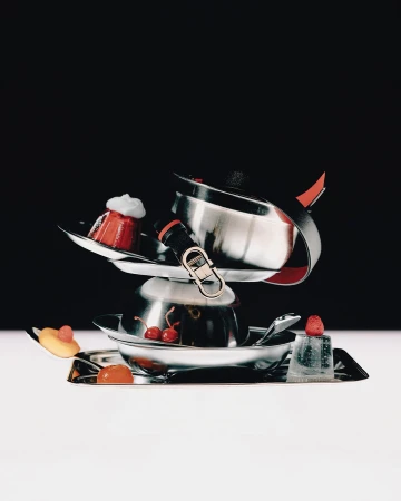 Stacked metallic cookware and trays garnished with desserts and cherries against a dark and light background