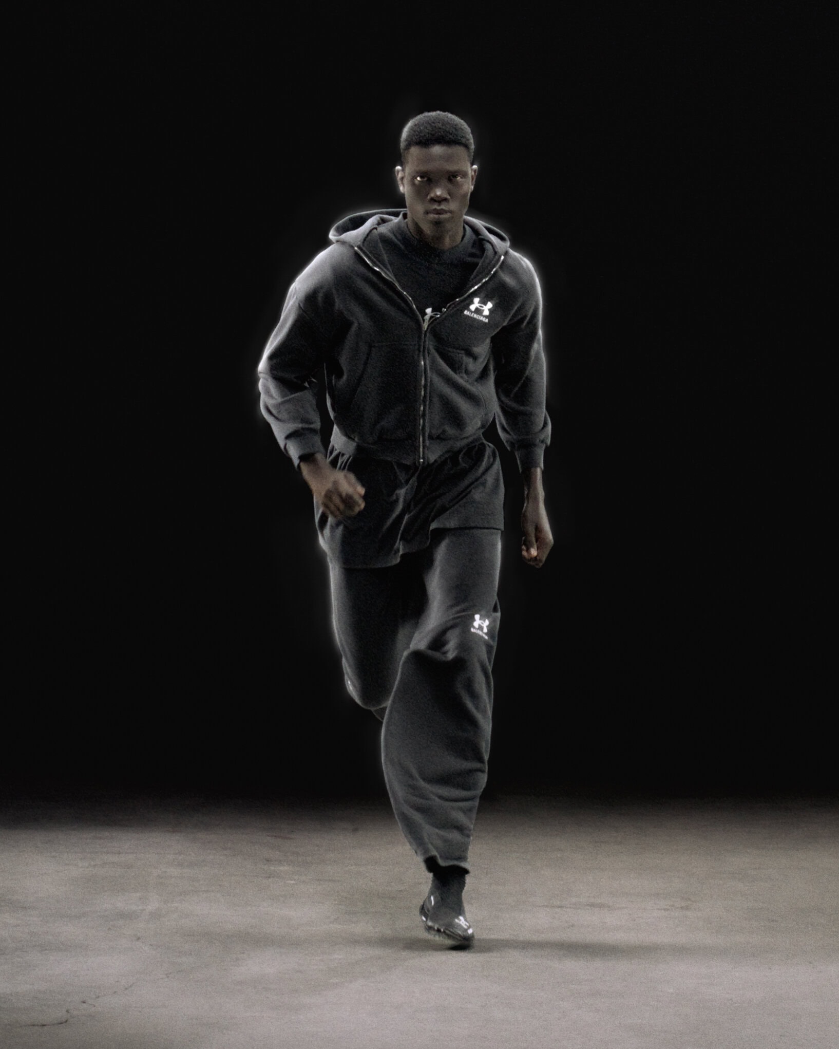 Black man running in a dark grey zip-up hoodie and sweatpants against a dark studio background