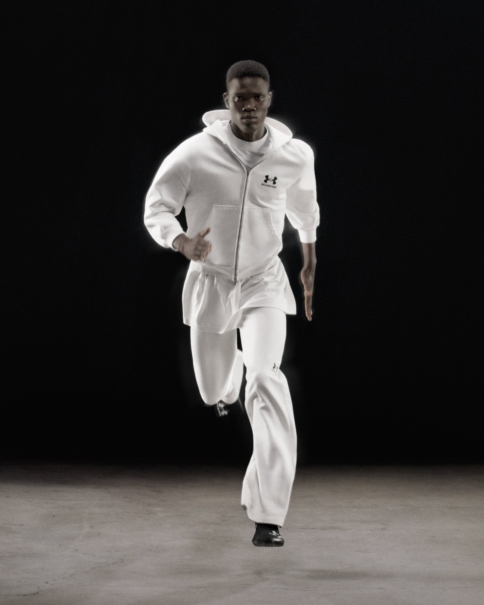 Black man running forward wearing a white hoodie and matching pants against a dark studio background