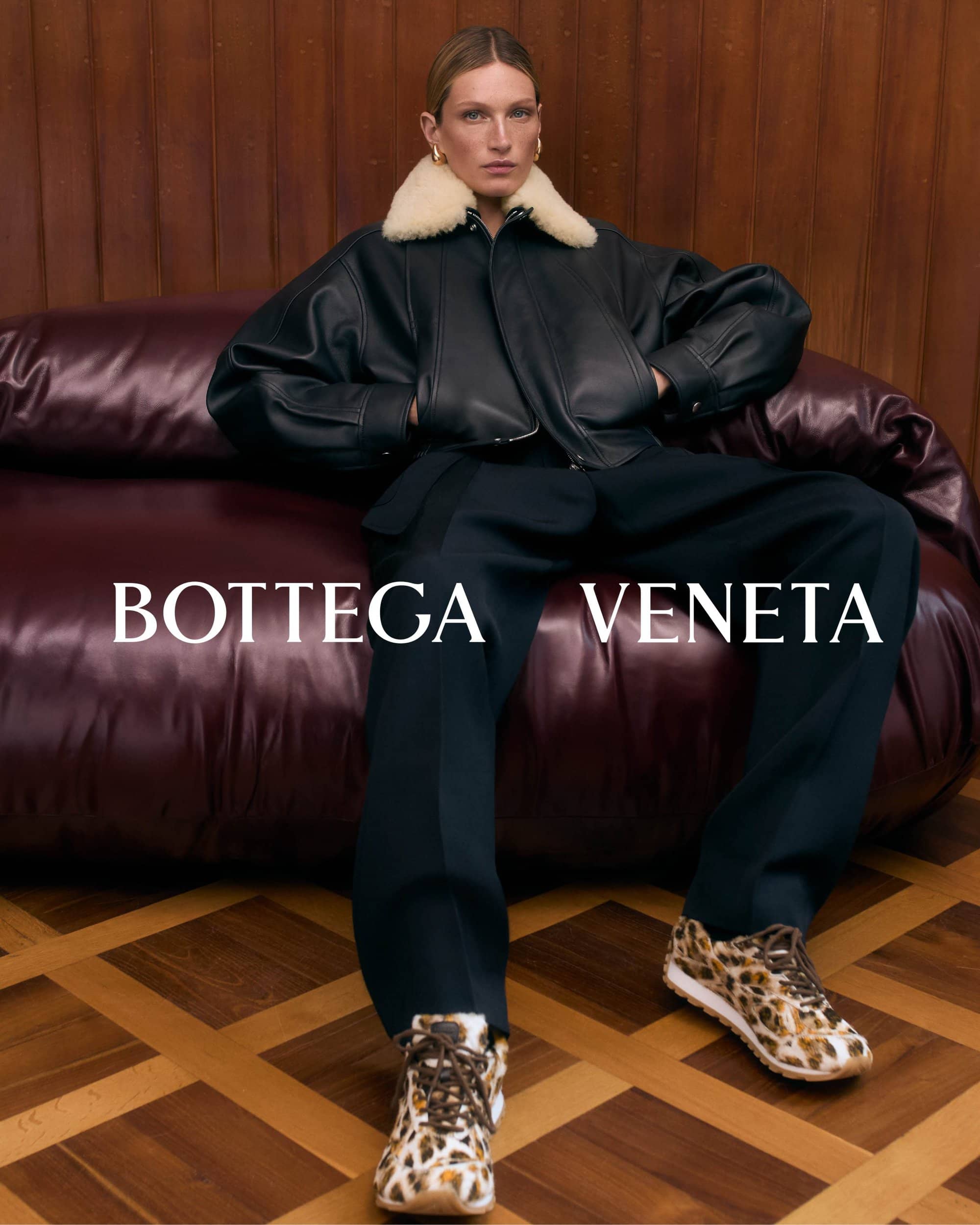 Model wearing a black leather jacket with a white collar sitting on a dark leather couch for Bottega Veneta