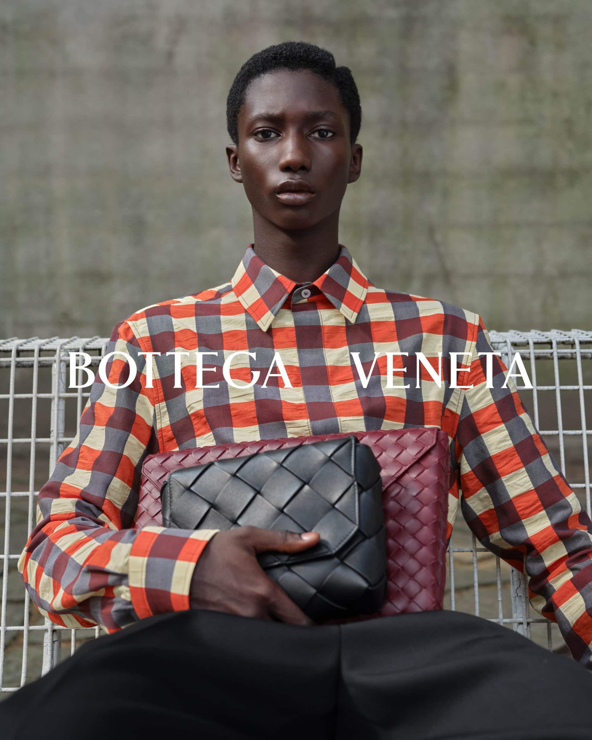 A Black man wearing a red plaid shirt holds a black woven leather clutch against an industrial wire mesh background.