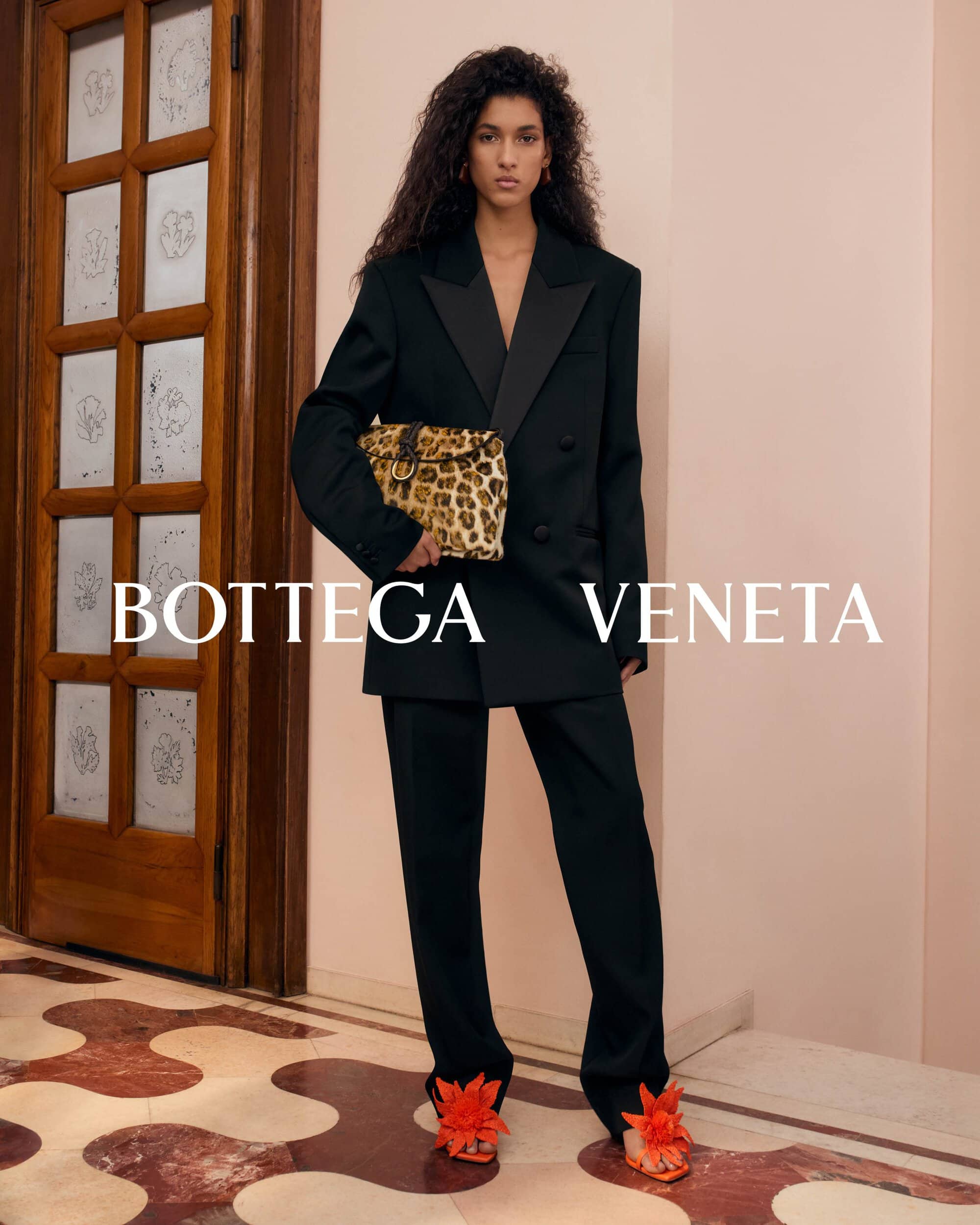 Model in a black suit holding a leopard print handbag standing inside a building with a tall wooden door and patterned floor.