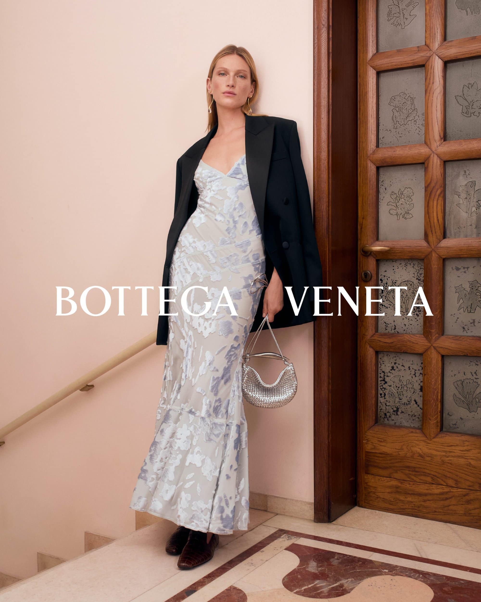 Model in a floral slip dress and black blazer leaning against a wooden doorframe on a staircase.