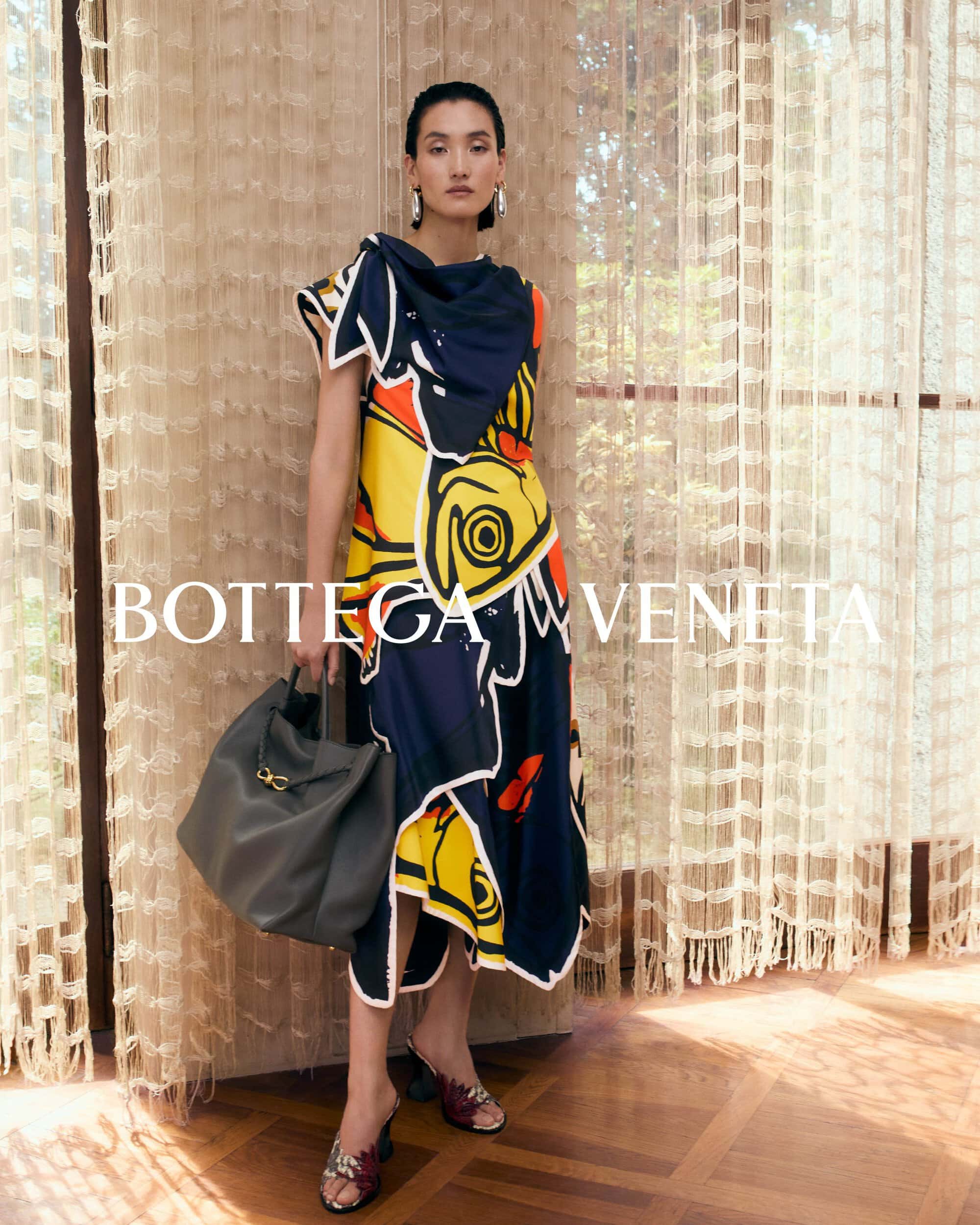 Model standing indoors wearing a colorful graphic dress and carrying a large dark handbag in front of textured curtains.