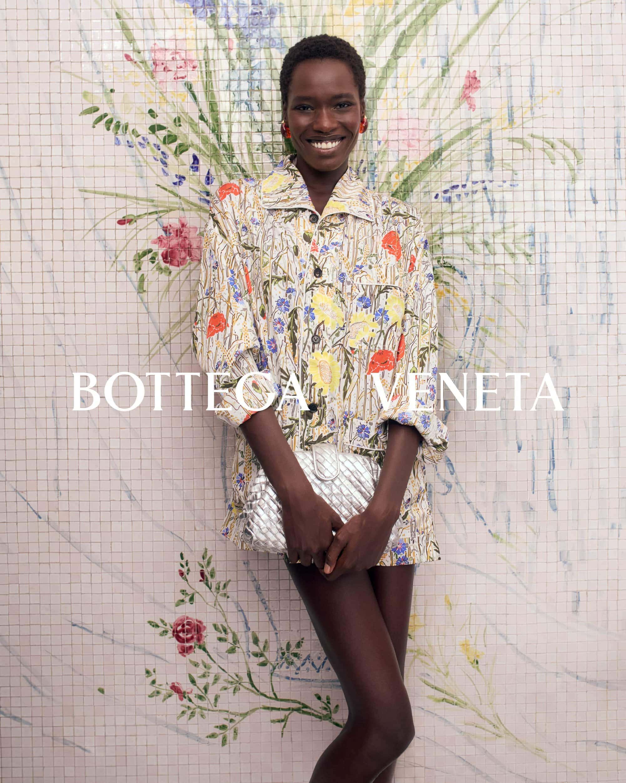 Smiling woman wearing a floral shirt and holding a metallic clutch against a tiled mosaic wall background