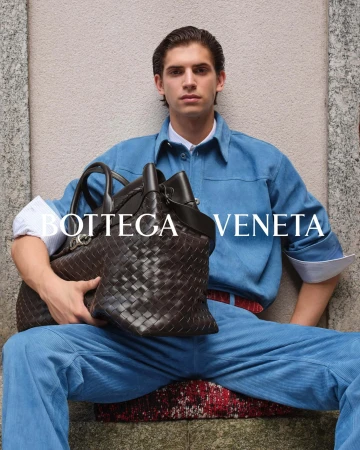 Man seated wearing a blue shirt and trousers holding a dark woven leather handbag against a stone building exterior