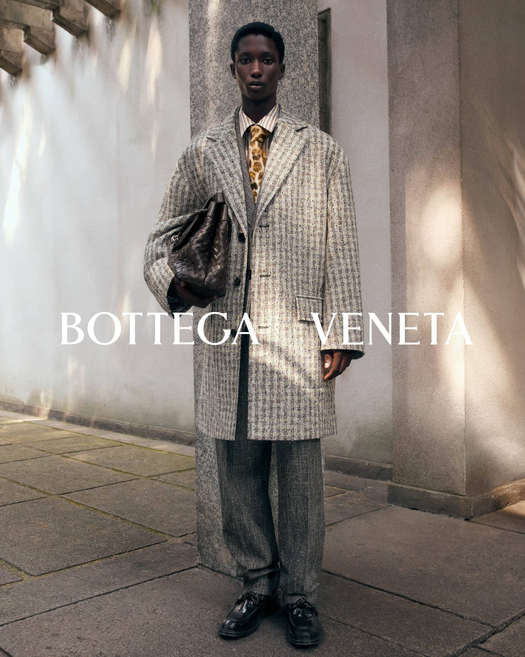 Black man standing outdoors wearing a grey patterned tweed coat holding a dark leather bag for Bottega Veneta Winter Solstice campaign