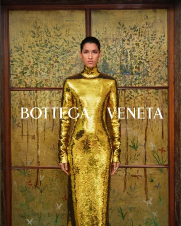 Model wearing a shimmering gold metallic turtleneck dress standing against an ornate painted decorative screen