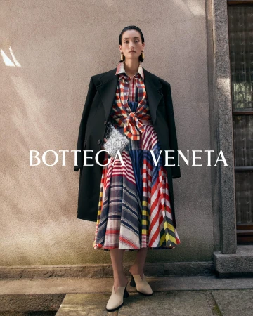 Model standing outdoors wearing a plaid skirt and black coat, holding a silver clutch against a stone building.