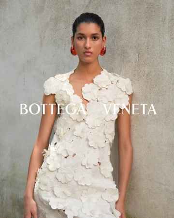 Model wearing a white floral appliqué dress and bold red earrings against a textured concrete wall for Bottega Veneta.