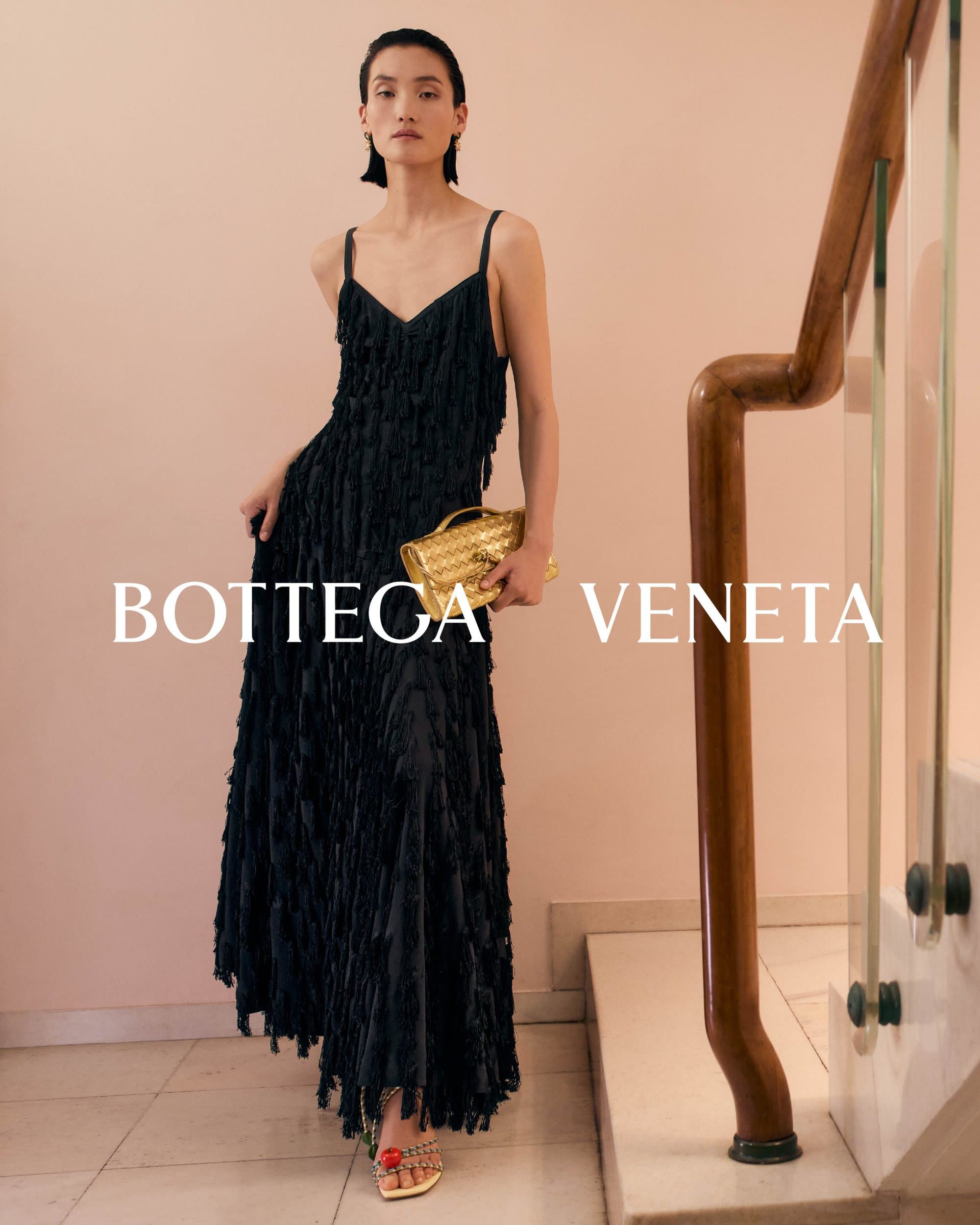 Model wearing a black fringed dress holding a gold clutch while standing on a staircase for Bottega Veneta