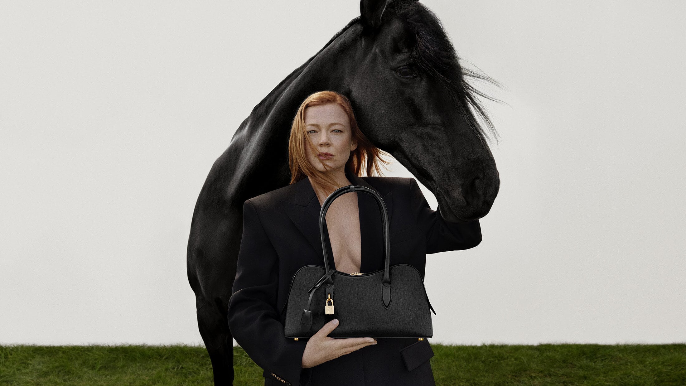 Sarah Snook in a black blazer holding a black handbag with a black horse standing behind her against a pale background