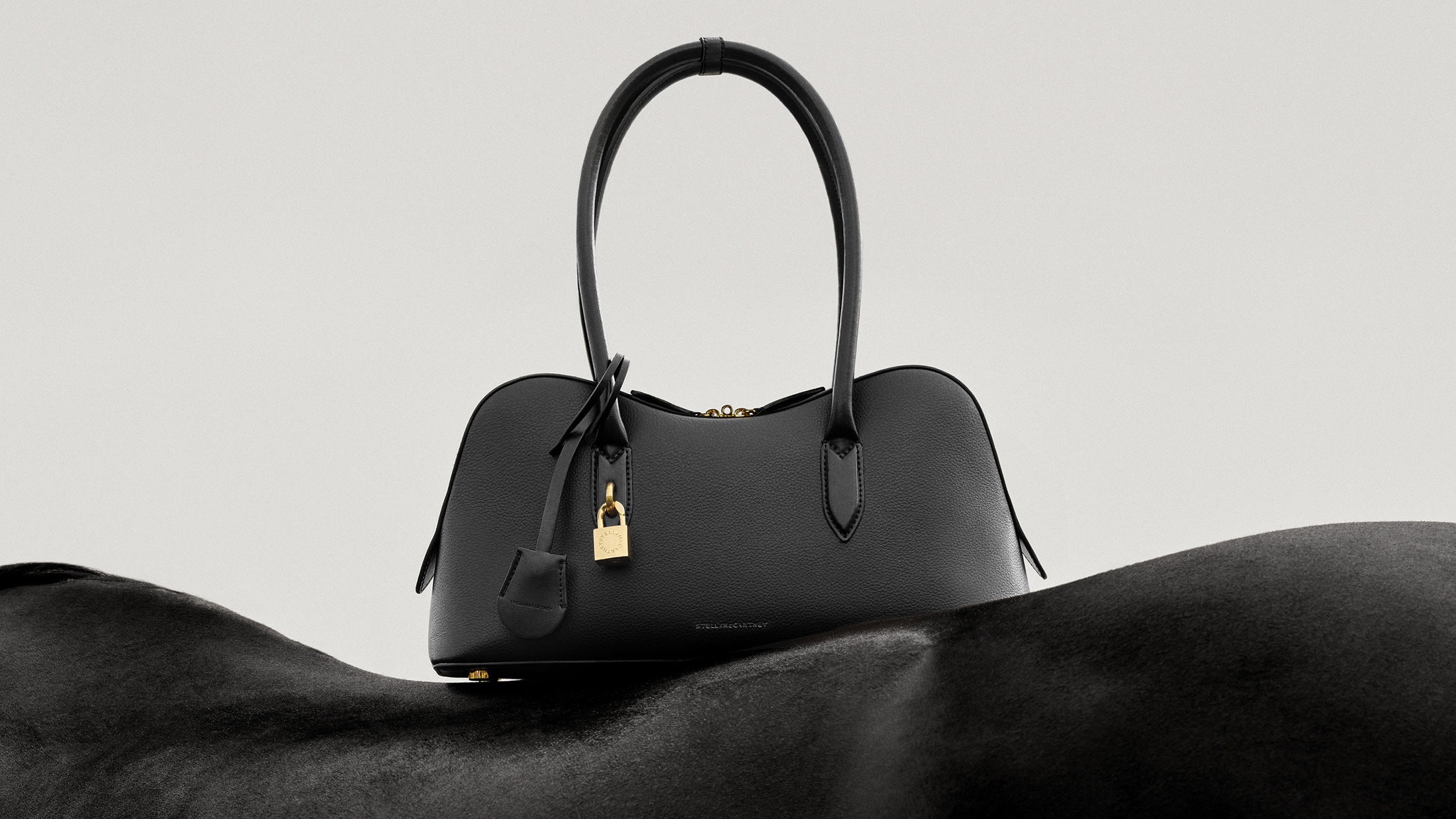 Black leather handbag with gold padlock resting on a black horse against a minimalist off-white seamless background.