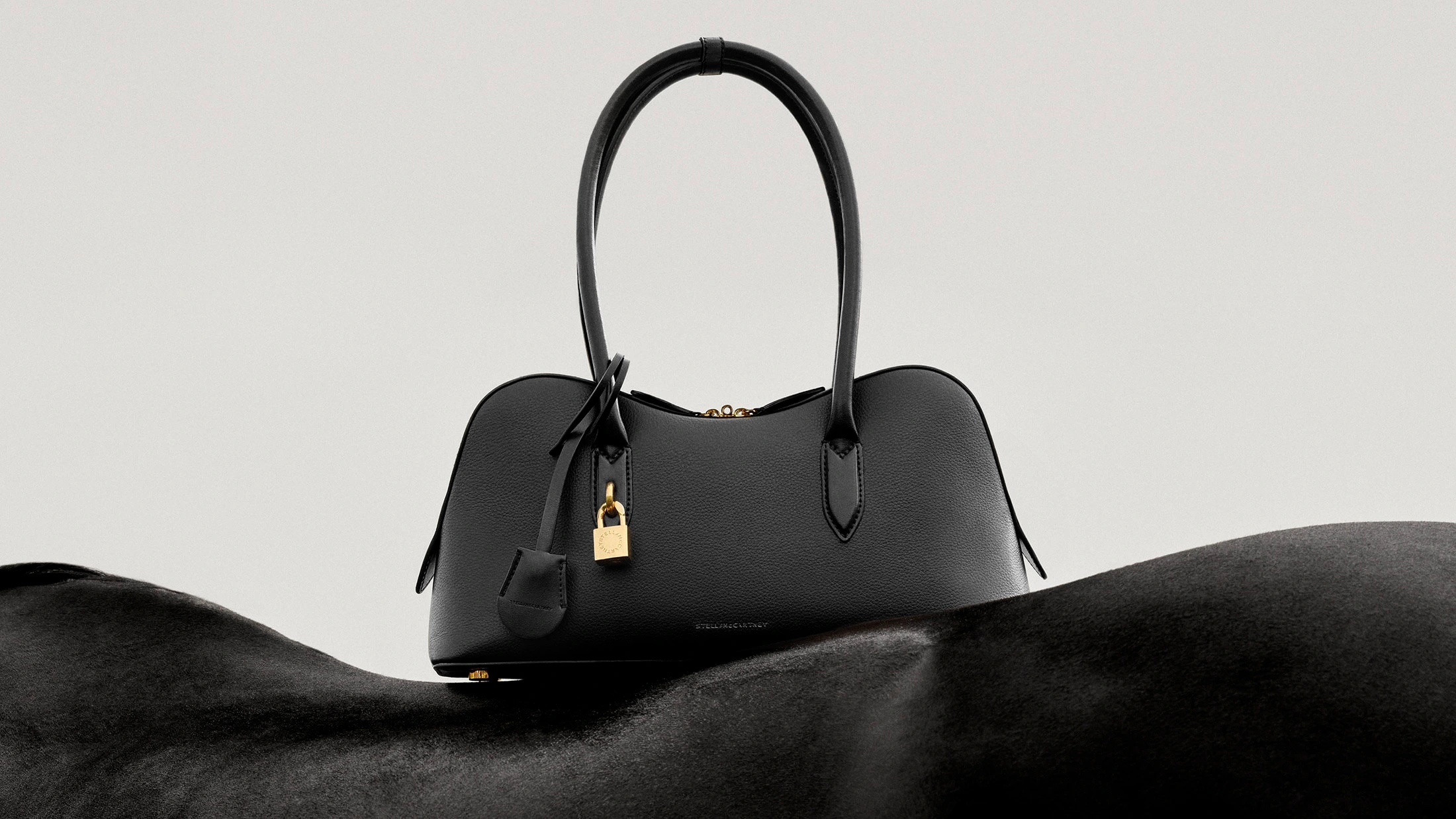 Black leather handbag with gold padlock resting on a black horse against a minimalist off-white seamless background.