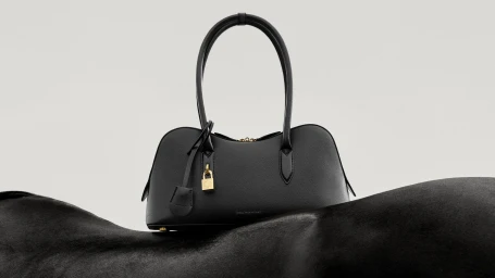 Black leather handbag with gold padlock resting on a black horse against a minimalist off-white seamless background.