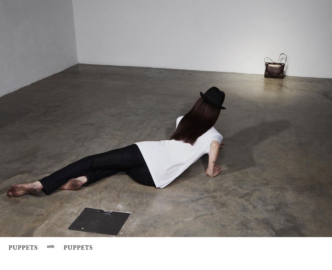 Person with long dark hair wearing a hat, white top, and black trousers lying on a concrete floor near a handbag.