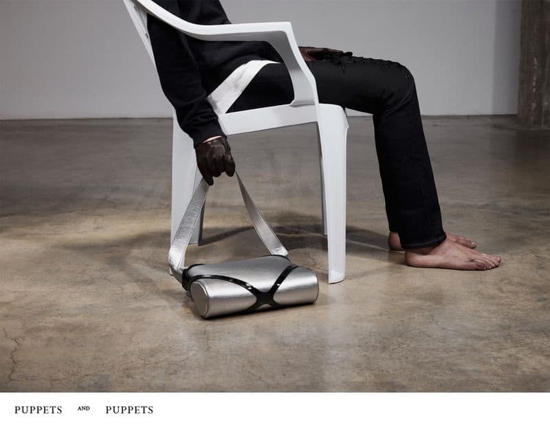 A person sitting barefoot on a white plastic chair holding a metallic silver handbag in a gloved hand.