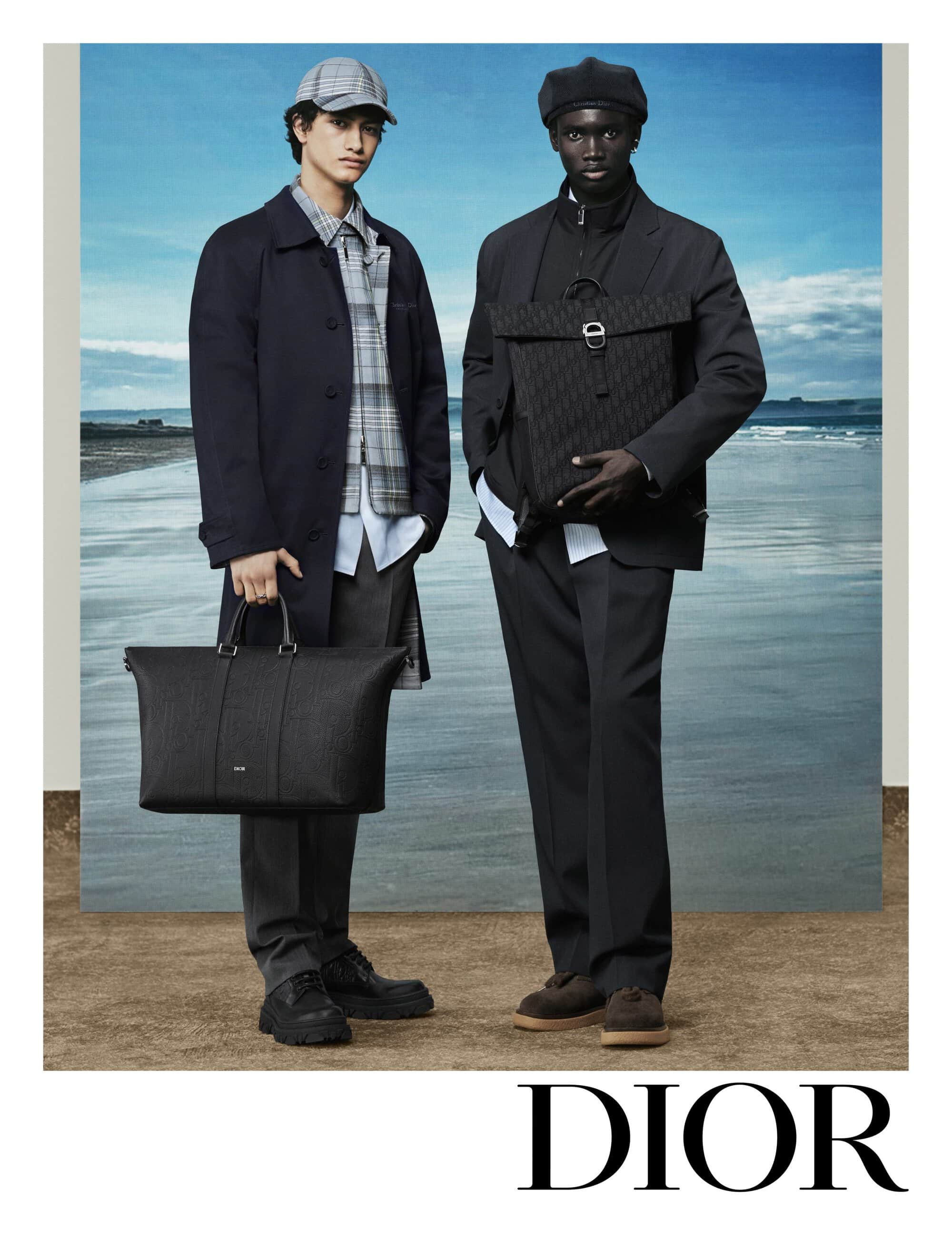 Two male models standing on a sandy ground holding black leather bags against a coastal backdrop for Dior.