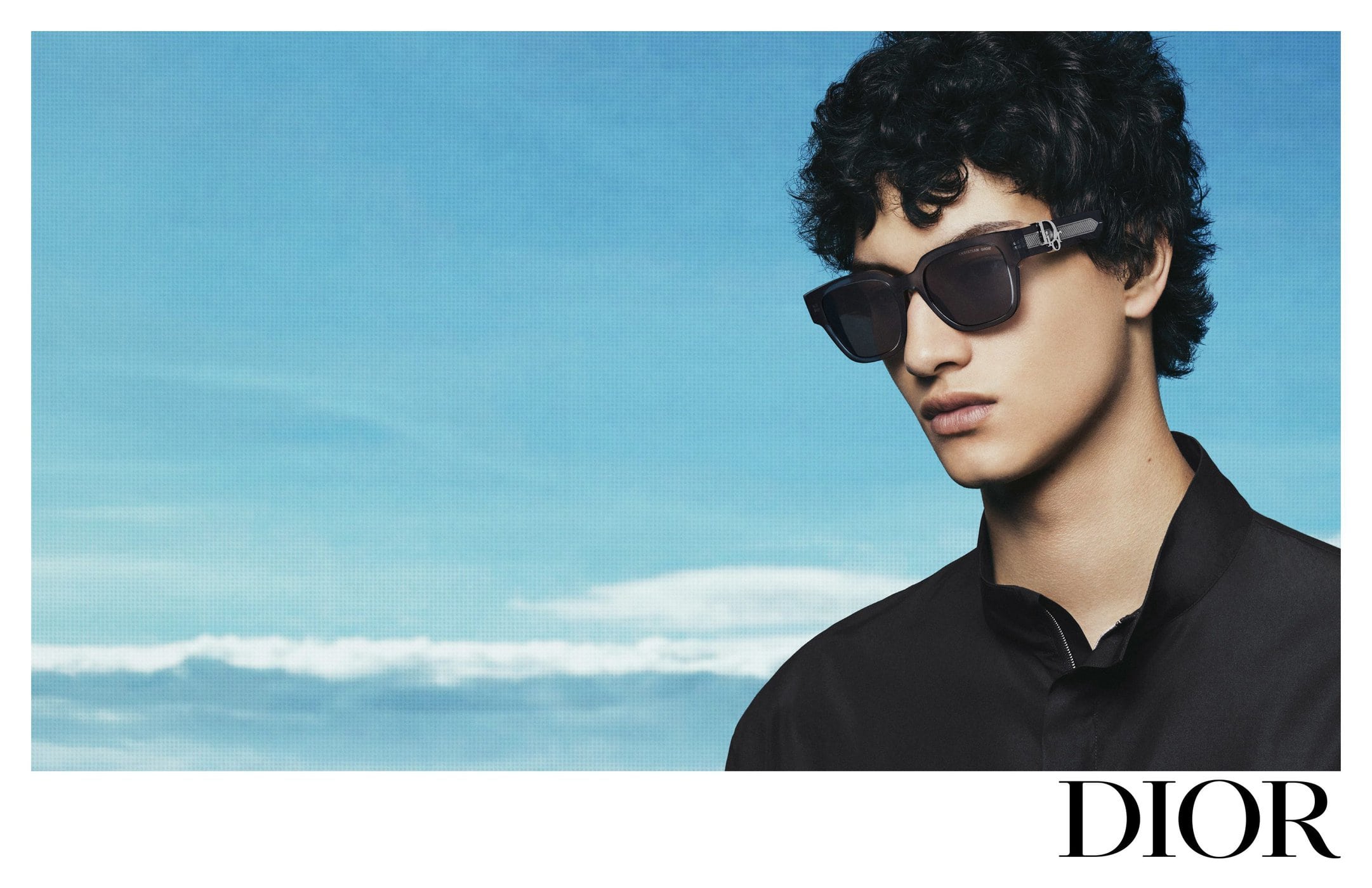 Close-up portrait of a male model wearing dark sunglasses and a black shirt against a blue sky backdrop with Dior logo