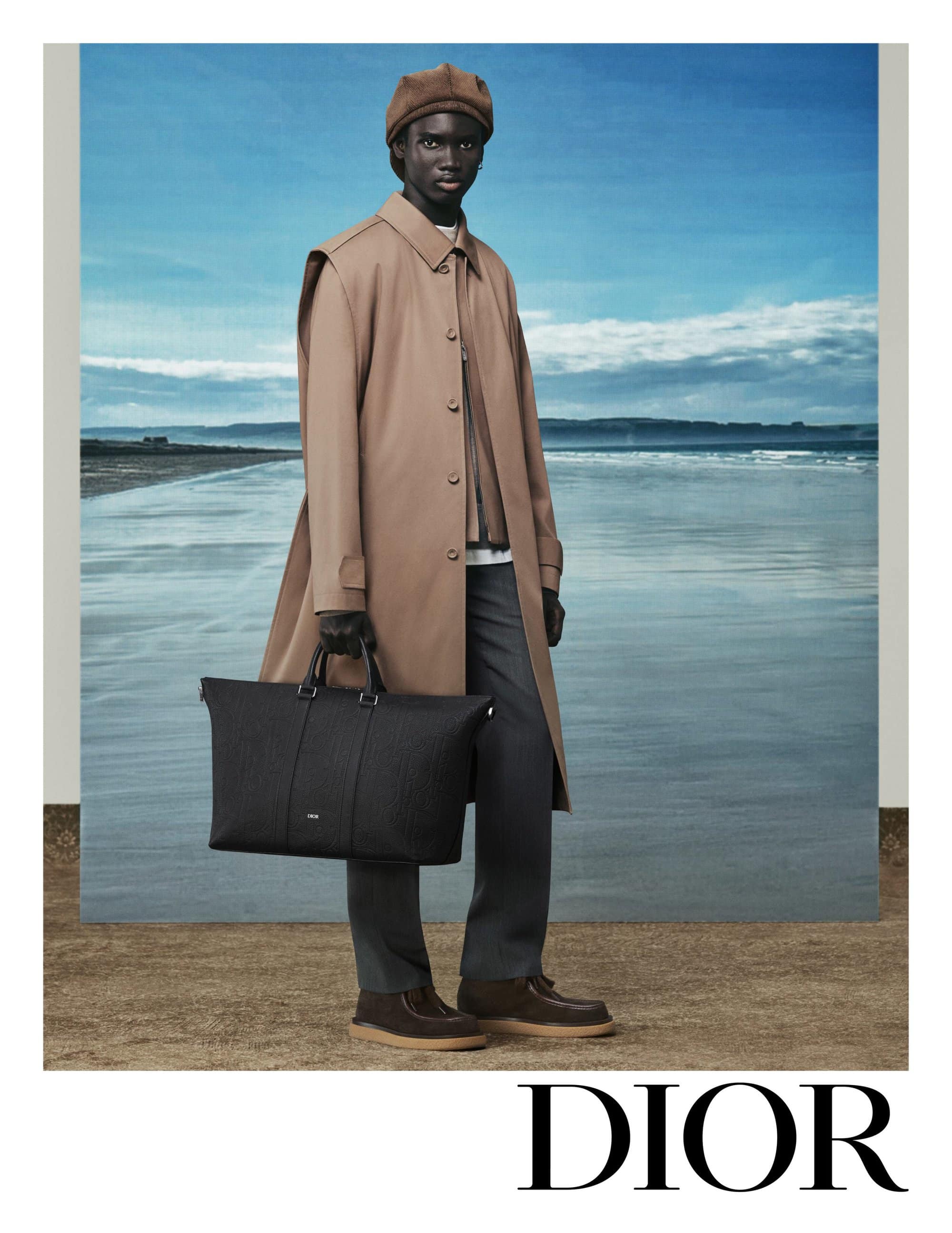 Model wearing a tan coat and cap holding a black leather bag against a backdrop of a sandy beach and blue sky for Dior.