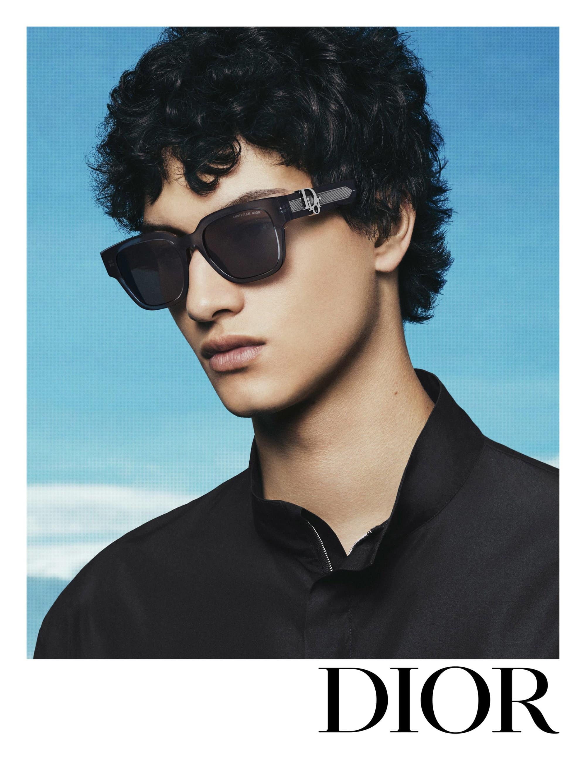 Model wearing dark sunglasses and a black shirt against a blue sky backdrop for Dior