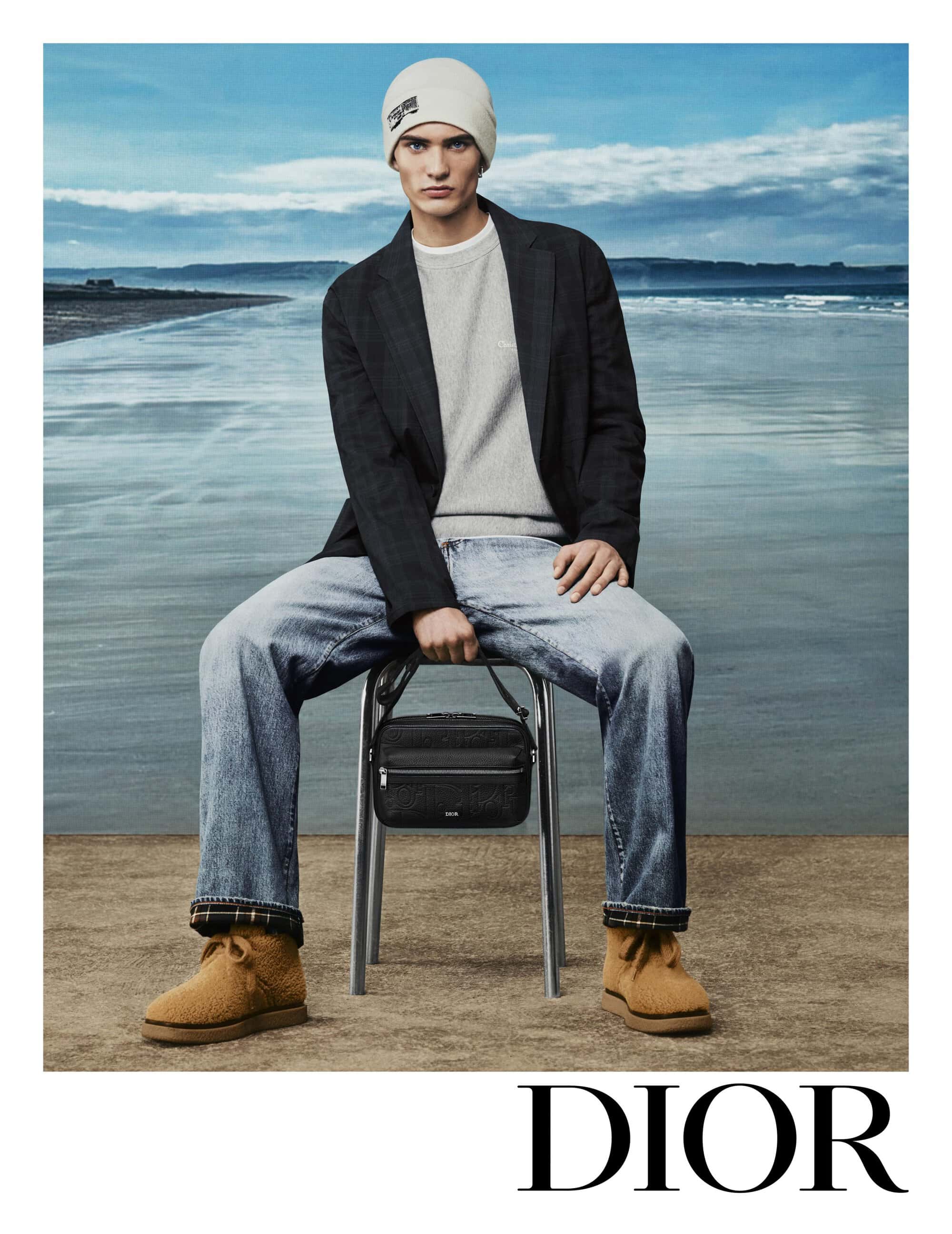 Model wearing a beanie, dark blazer, and light jeans seated on a stool holding a black handbag against a coastal backdrop.