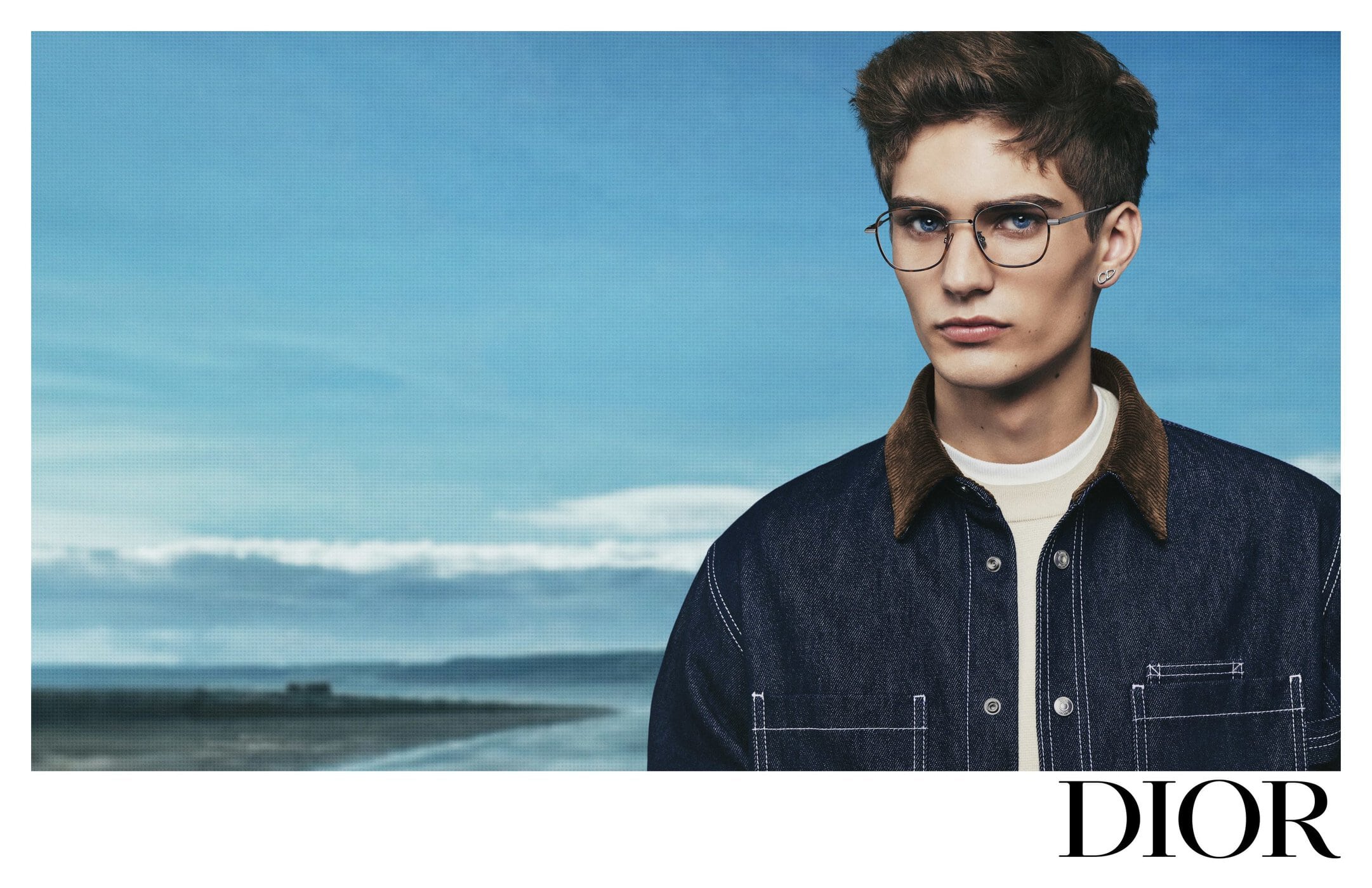 Close-up portrait of a young man wearing wire-frame glasses and a dark denim jacket against a blue sky background.