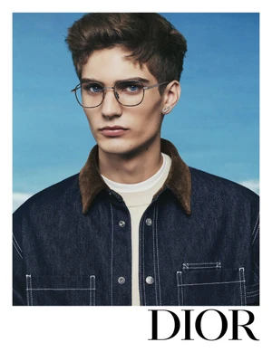 Close-up portrait of a young man wearing wire-rimmed glasses and a dark denim jacket against a blue sky backdrop for Dior