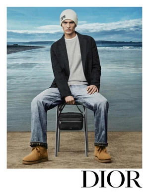 Model wearing a beanie, dark blazer, and light jeans seated on a stool holding a black handbag against a coastal backdrop.