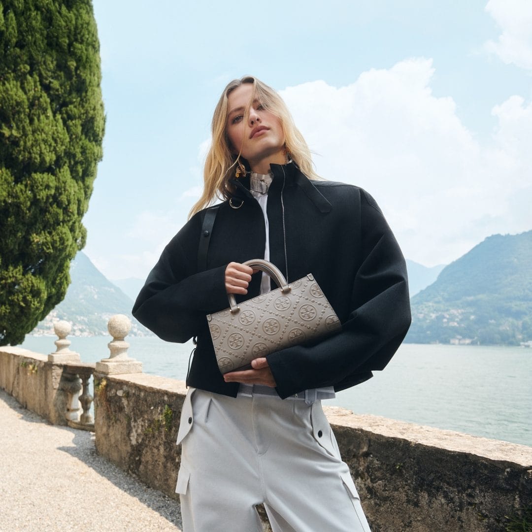 Model in black jacket holding a patterned handbag by a lakeside stone balustrade with mountains in the background
