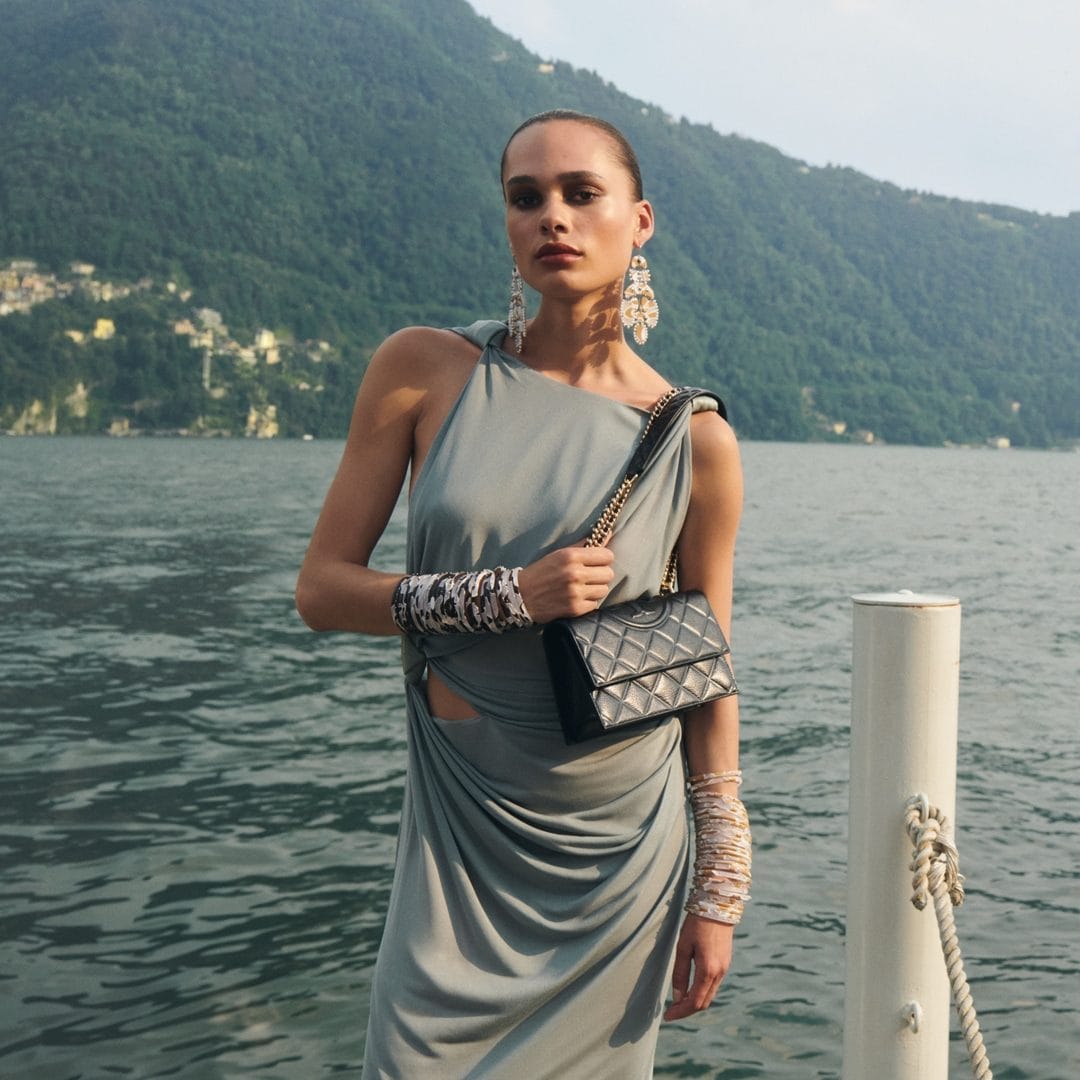 Model in grey draped dress holding a black quilted handbag against Lake Como, Italy, for Tory Burch Holiday 2024.