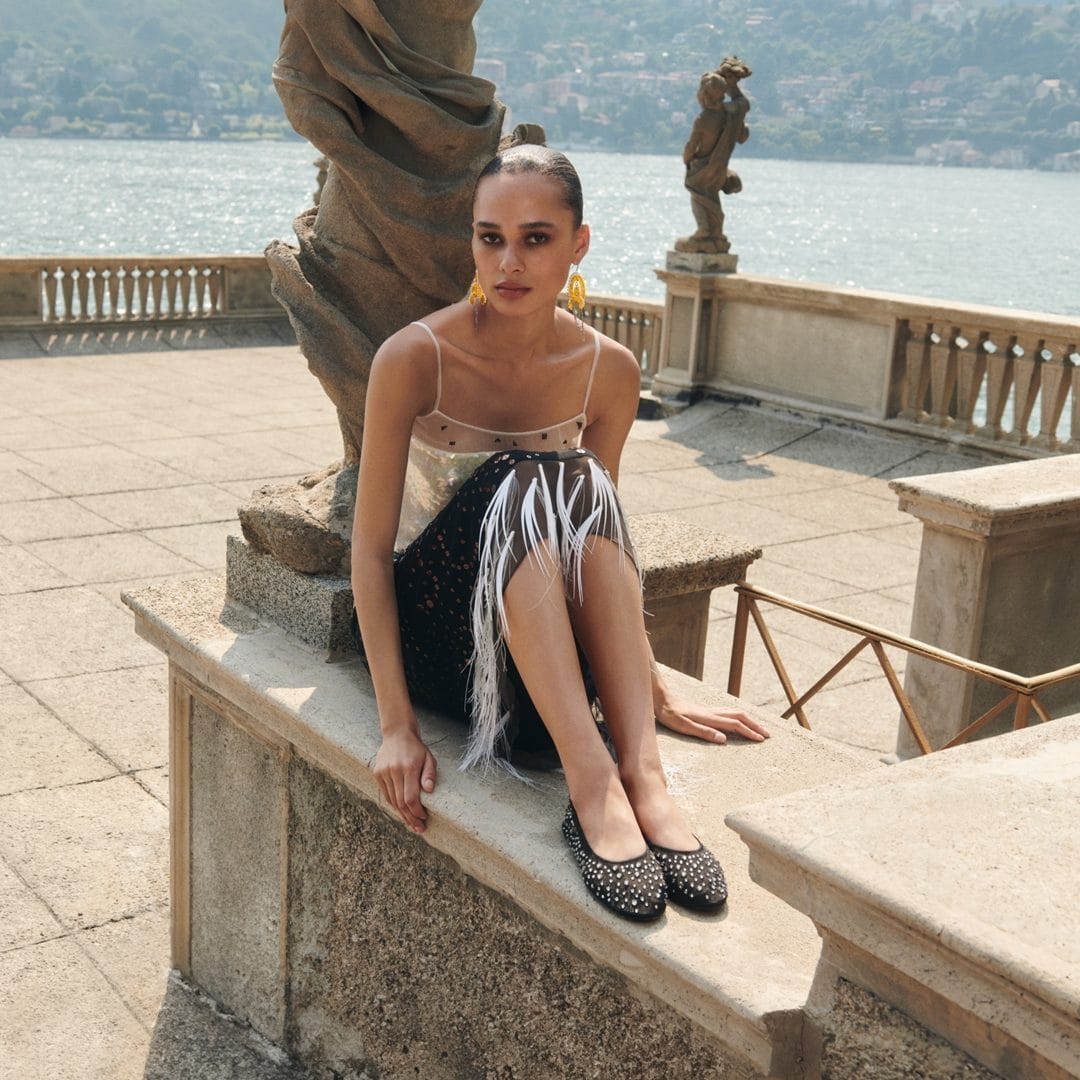Model seated on a stone balustrade by a lake wearing a feathered skirt and embellished flats in Lake Como