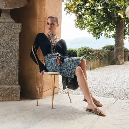 Model seated on a gold wire chair outdoors wearing a black long-sleeve top and patterned blue skirt