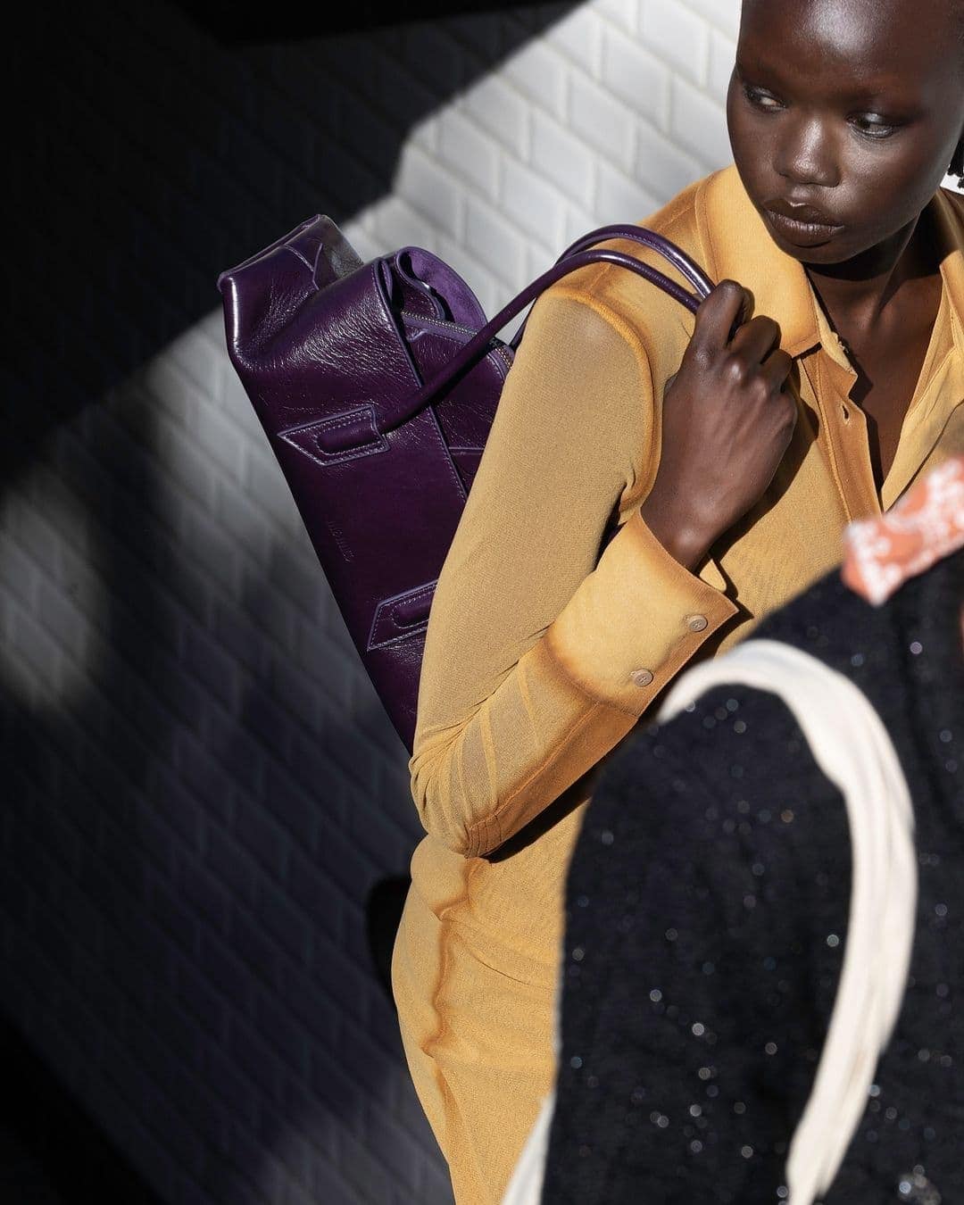Model in a yellow top holding a purple leather handbag against a tiled background for The Attico Fall 2024 campaign