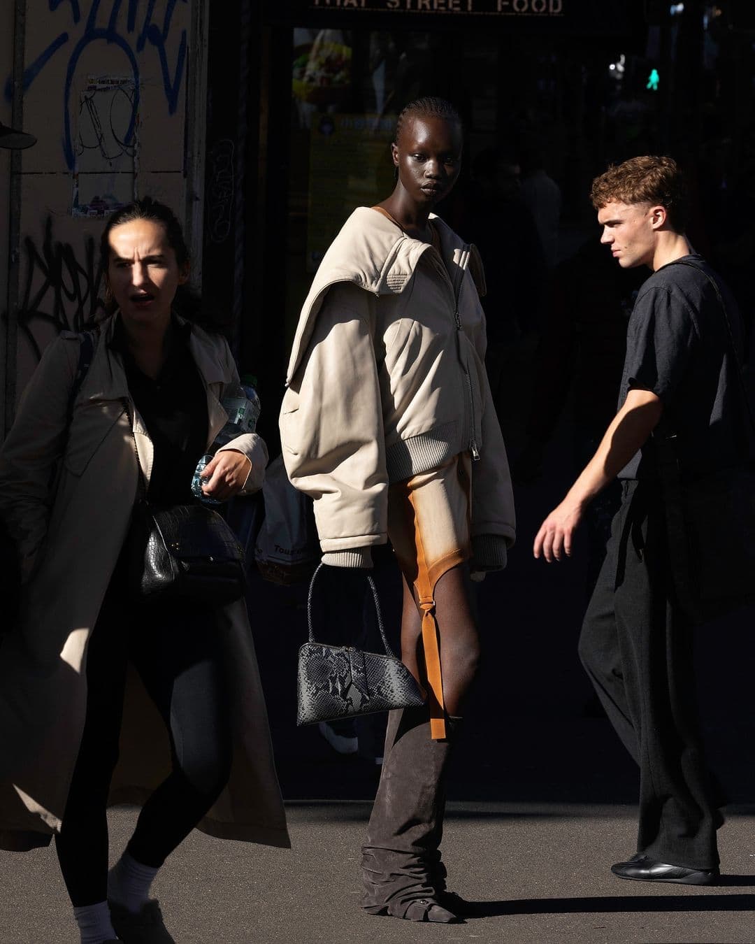 Three people walking on an urban street, featuring a model in a beige jacket and holding a patterned handbag.