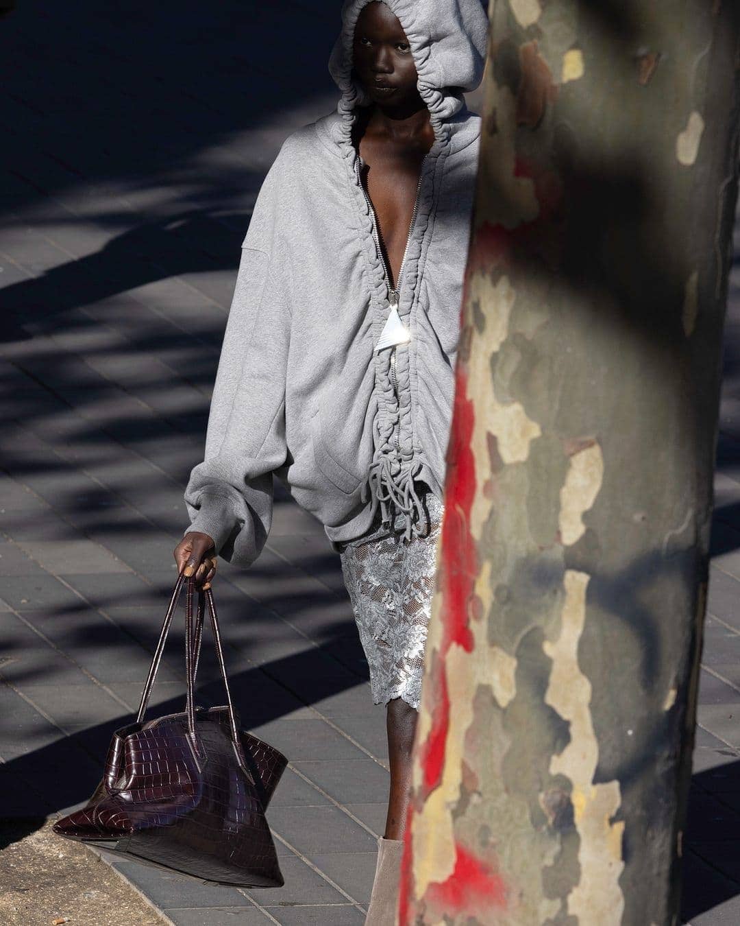 Model in grey hooded sweatshirt holding dark brown leather handbag outdoors on paved surface