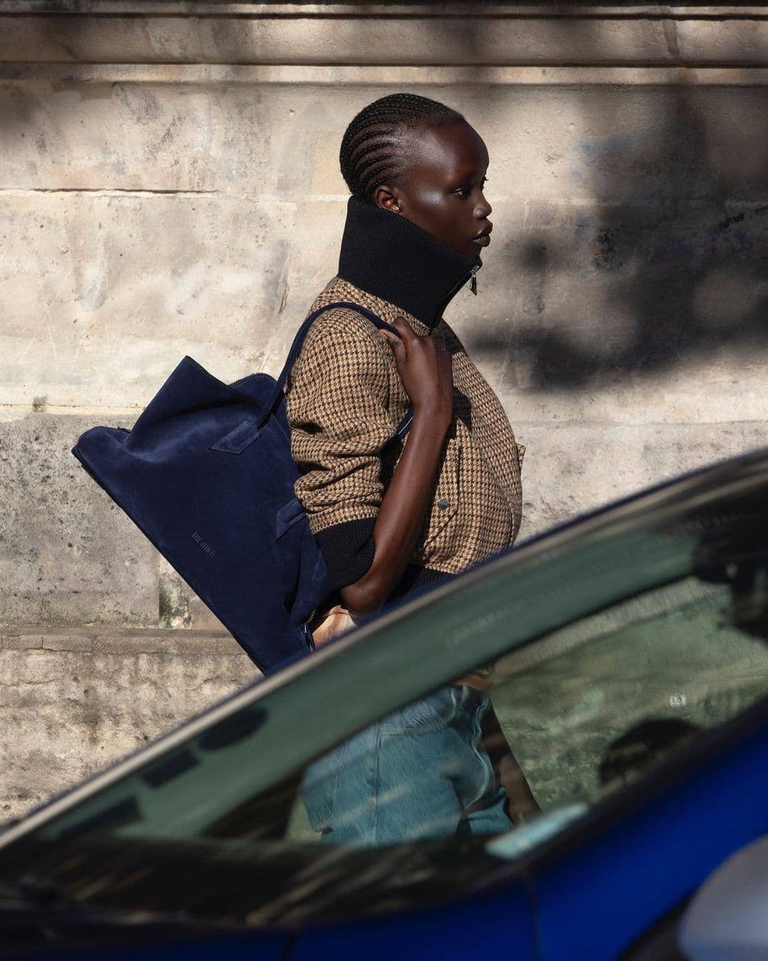 Model in patterned jacket with high collar carrying a blue tote bag outdoors near a car