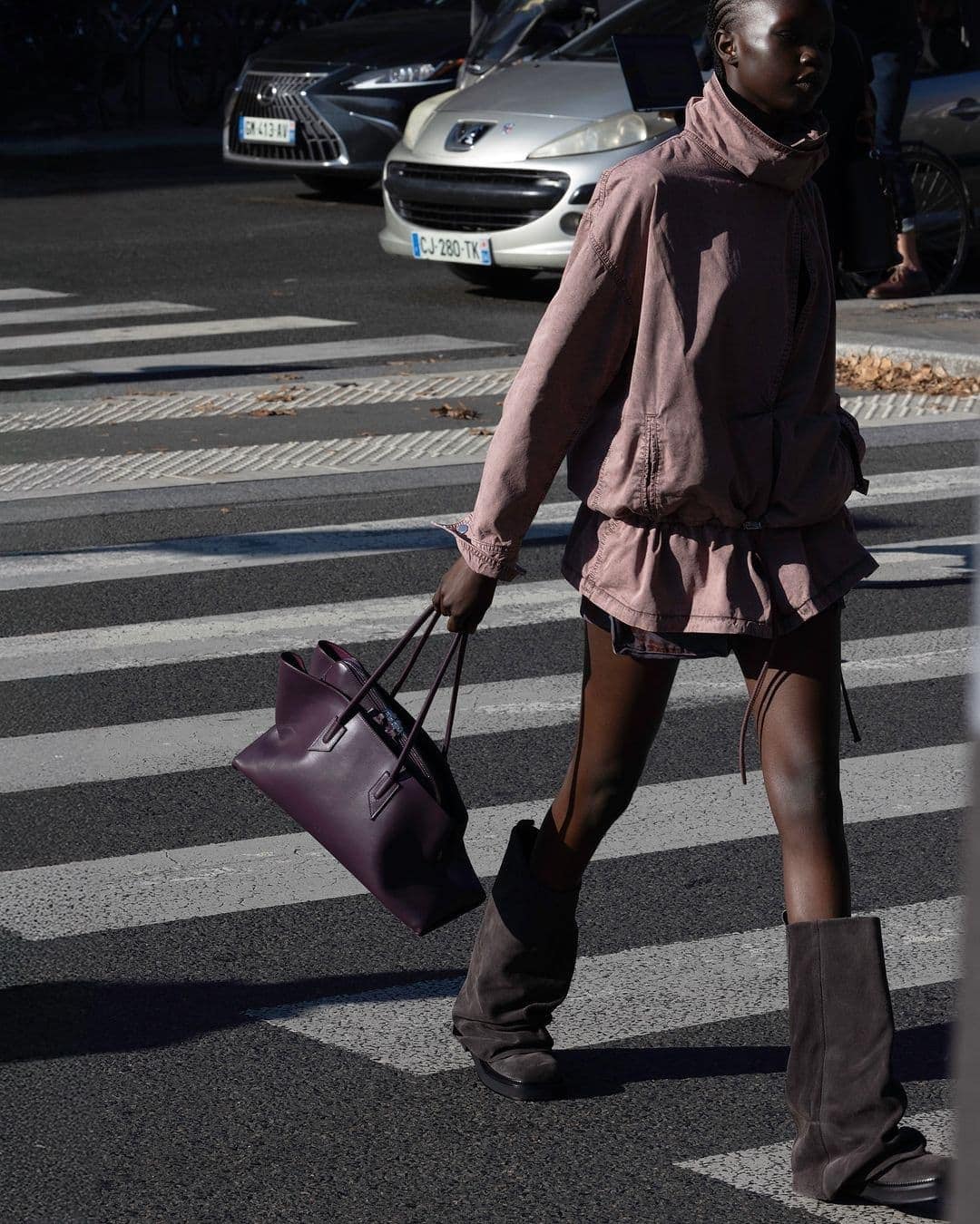 Black woman walking across a city street holding a purple handbag wearing a pink jacket and grey boots.