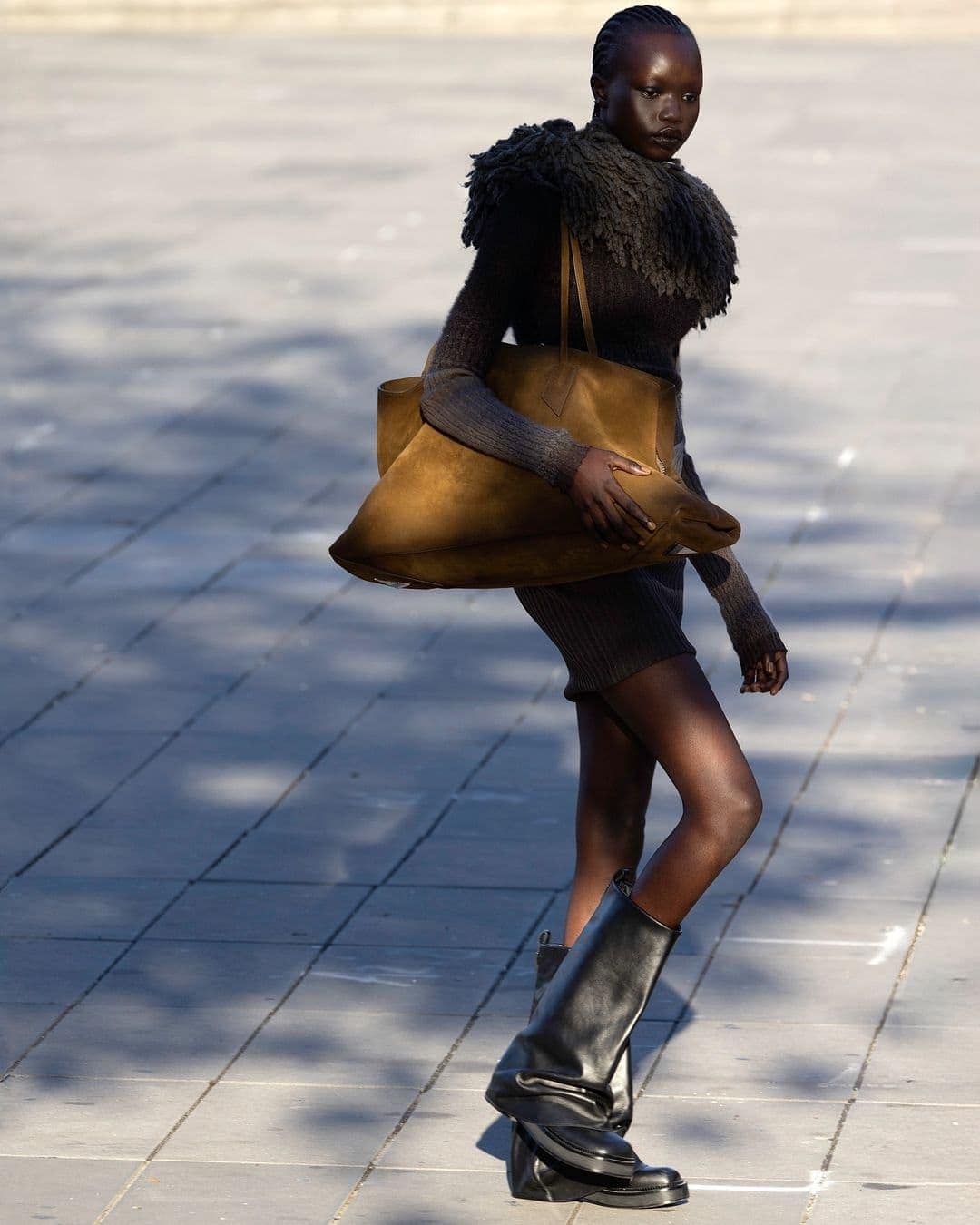 Model wearing dark knitwear and tall black boots holding a large brown suede handbag outdoors on paved stone ground