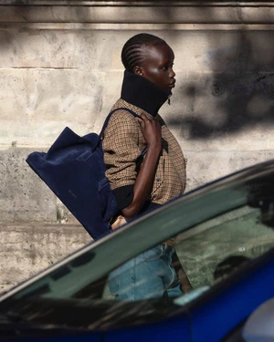 Model in patterned jacket with high collar carrying a blue tote bag outdoors near a car