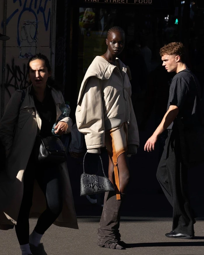 Three people walking on an urban street, featuring a model in a beige jacket and holding a patterned handbag.