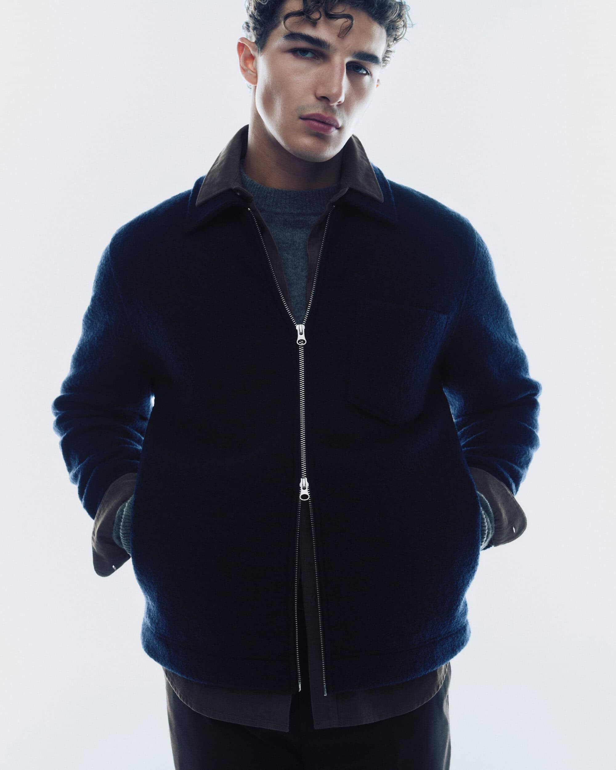 Model in a dark navy blue zip jacket and grey sweater against a plain white studio background for Samsoe Samsoe.