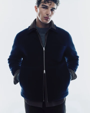 Model in a dark navy blue zip jacket and grey sweater against a plain white studio background for Samsoe Samsoe.