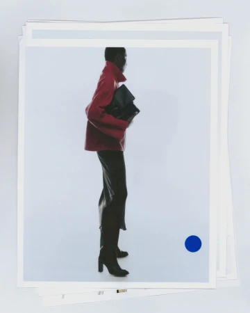 Model wearing a red jacket and black skirt holding a dark handbag against a plain white studio backdrop.