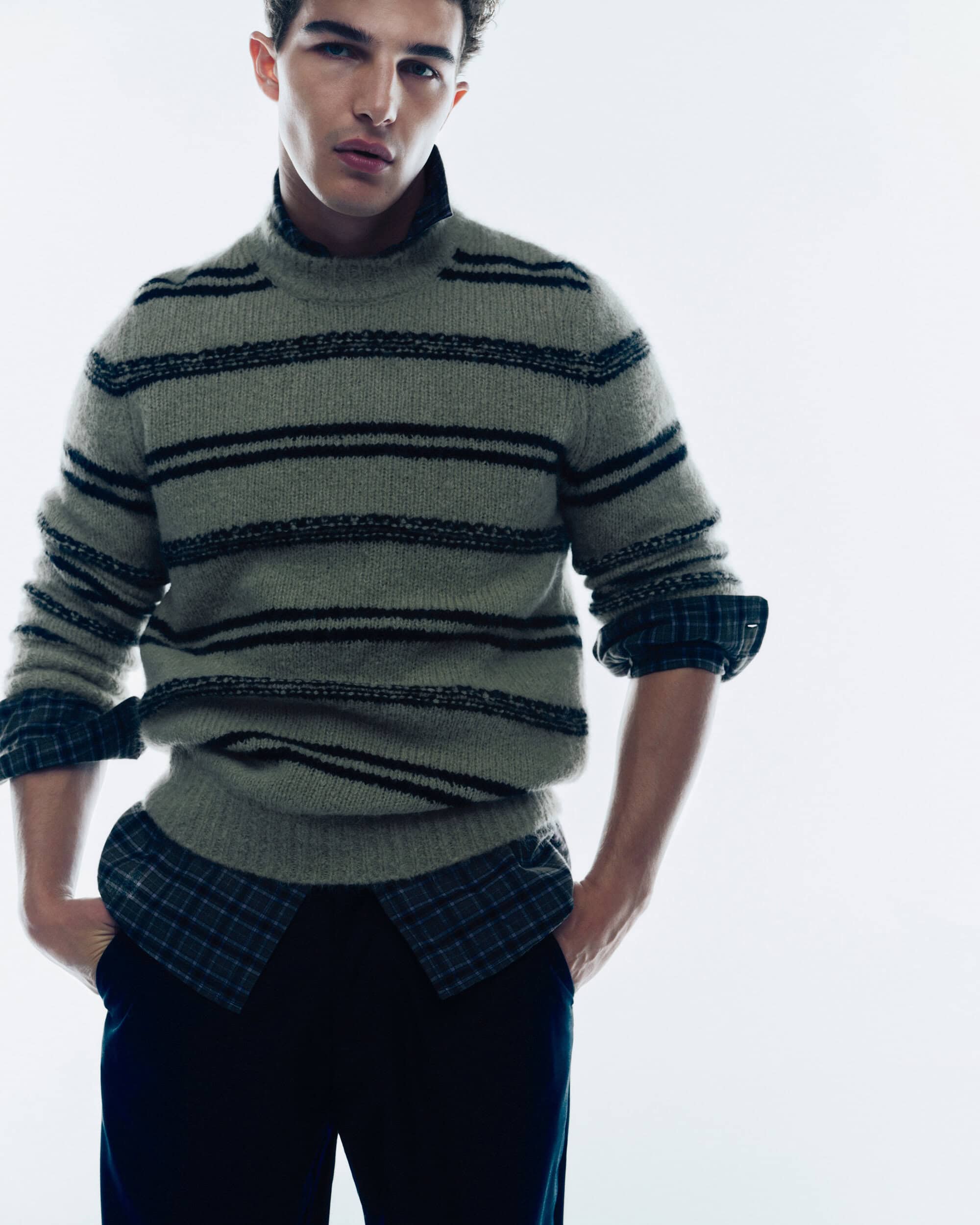 Male model wearing a striped knit sweater over a plaid shirt against a white seamless background